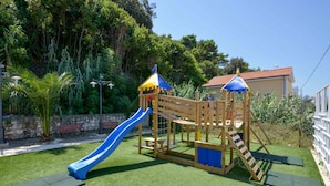 Ein hölzerner Spielplatz mit einer blauen Rutsche und Klettergerüsten, umgeben von grünem Gras und Bäumen.