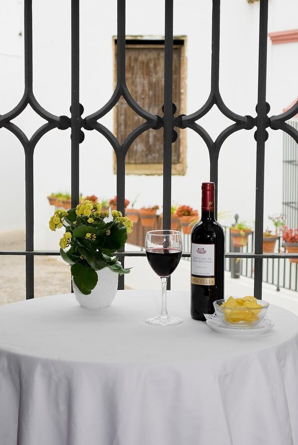 A table with a bottle of wine, a glass, and a bowl of fruit on a balcony with a metal railing and potted plants in the background.
