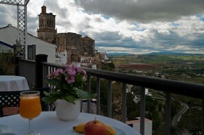 A balcony with a table set for a meal, a drink, and a fruit plate, overlooking a historic building and a scenic landscape.