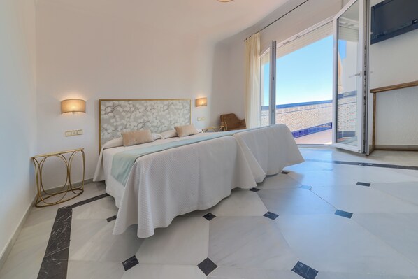 A bedroom with a bed, a nightstand, a lamp, and a view of the beach through a large window.