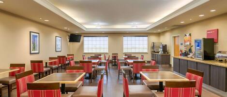 A hotel dining area with multiple tables and chairs, a kitchen counter, and framed pictures on the walls.