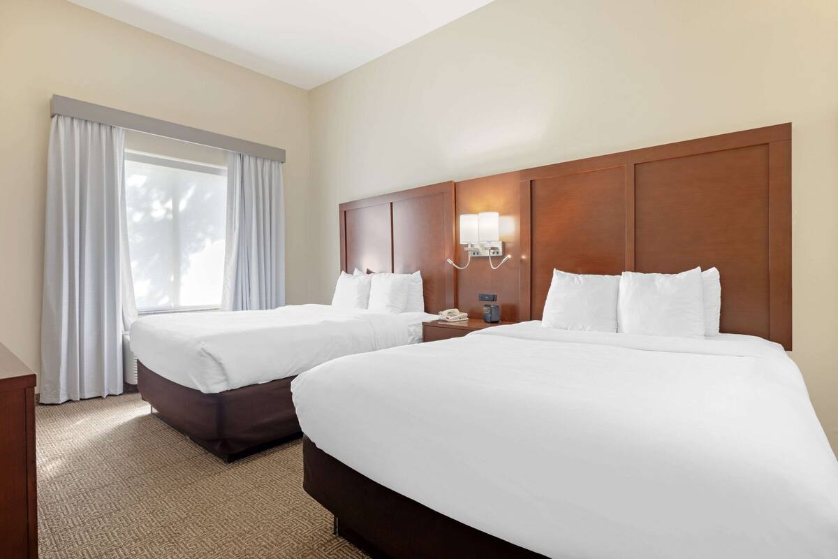 A hotel room with two beds, a large window with curtains, a wooden headboard, and a bedside table with a lamp and phone.