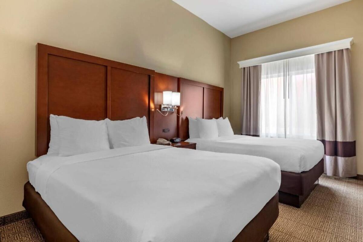 A hotel room with two beds, a wooden headboard, a nightstand with a lamp, and a window with curtains.