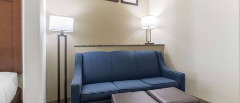 A hotel room with a blue sofa, a brown ottoman, a standing floor lamp, and a bed with white bedding.