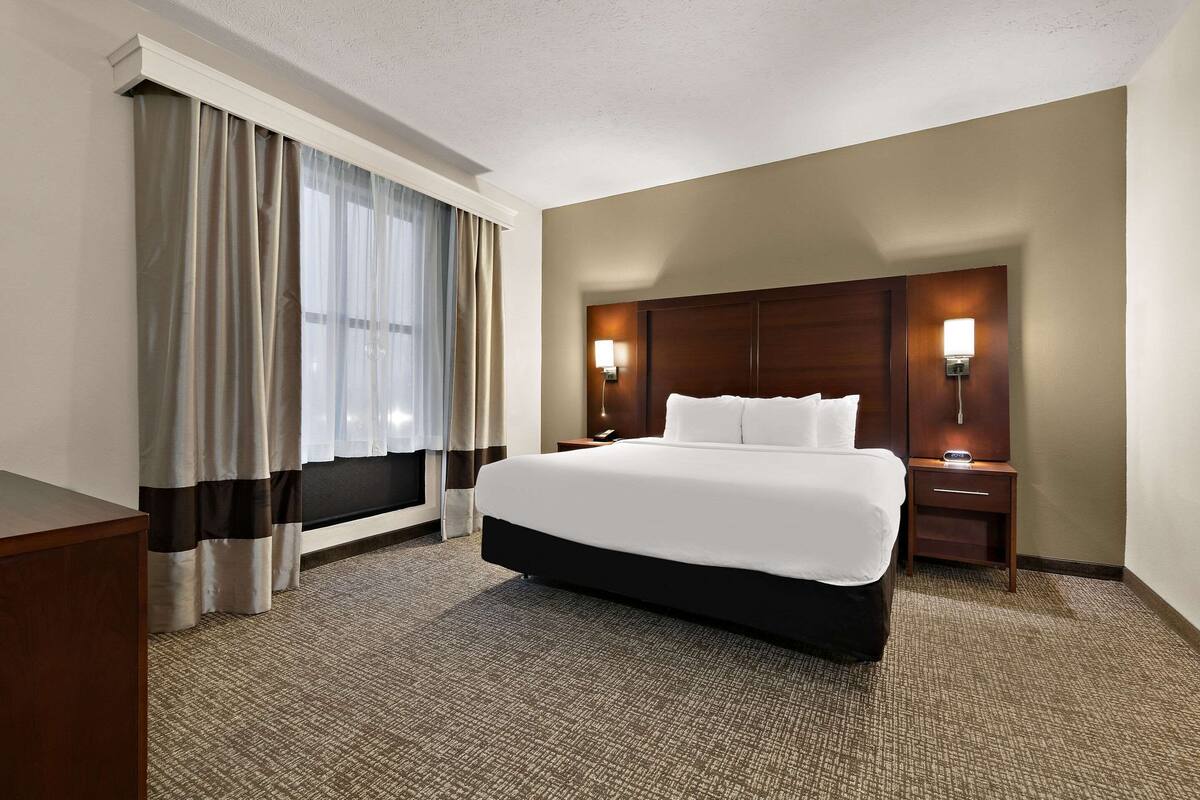 Een hotelkamer met een groot bed, een nachtkastje met lamp en een raam met gordijnen.
