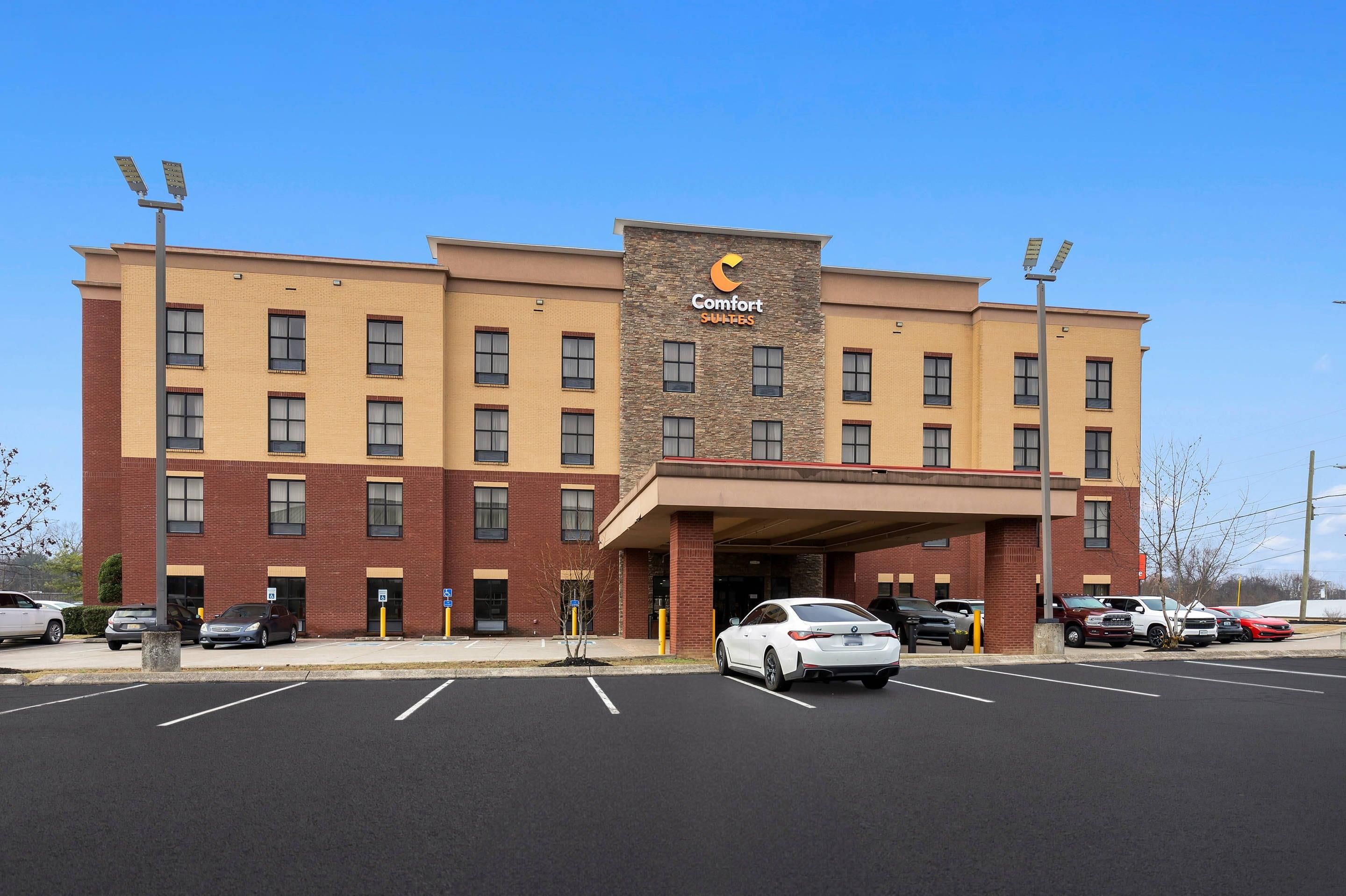 Un hotel de varios pisos con entrada central, estacionamiento y un cartel que indica 'Comfort Suites'.