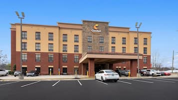 Un hotel de varios pisos con entrada central, estacionamiento y un cartel que indica 'Comfort Suites'.