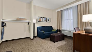 A hotel room with a blue sofa, a wooden desk, a lamp, a small table, a wall-mounted shelf, and an air conditioning unit.