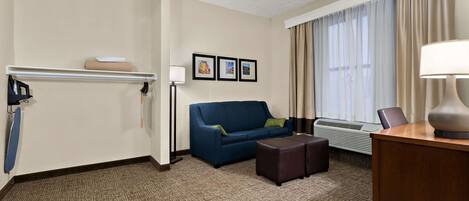 Habitación de hotel con sofá azul, escritorio de madera, lámpara, mesita pequeña, estante montado en la pared y unidad de aire acondicionado.