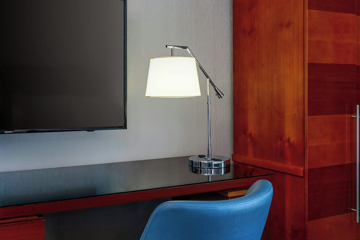 Habitación de hotel moderna con televisor de pantalla plana, un escritorio con lámpara y una silla azul.