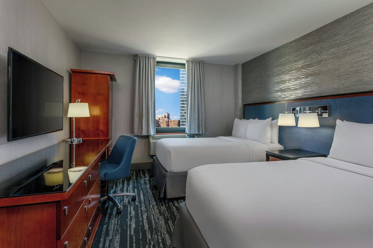 Habitación de hotel con una cama grande, un escritorio con televisor, una silla, una lámpara de escritorio y vistas a la ciudad a través de la ventana.