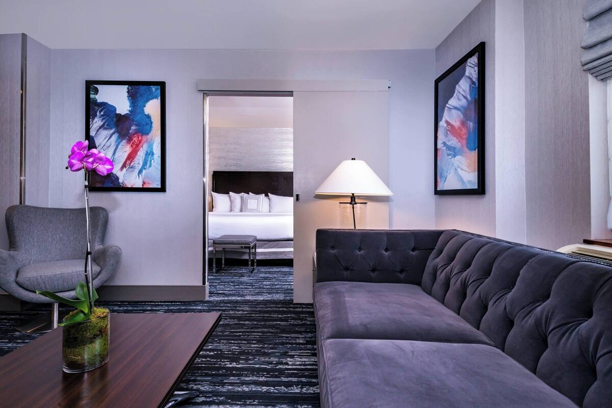 Una habitación de hotel moderna con un sofá gris, un sillón gris, una mesita con un jarrón de flores, una cama con ropa blanca y arte abstracto en las paredes.