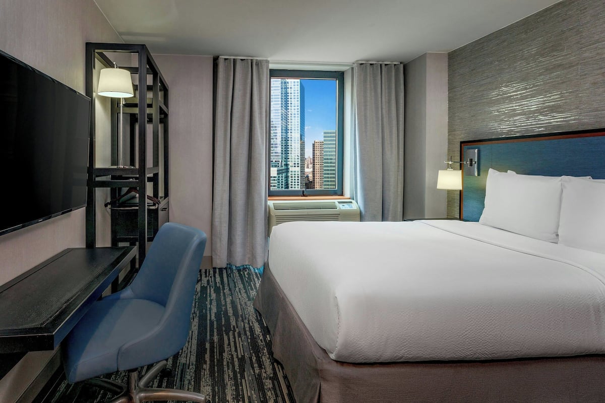 Habitación de hotel con cama, escritorio, silla, televisor y ventana con vista a la ciudad.