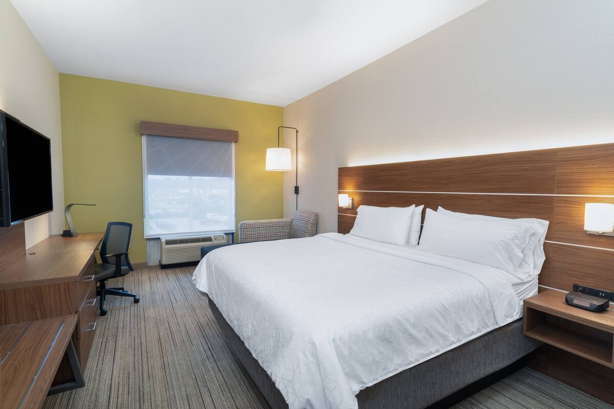 Une chambre d’hôtel avec un grand lit, un bureau avec une chaise, une télévision et une fenêtre avec des stores.