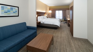 A hotel room with a bed, a blue sofa, a wooden coffee table, a framed picture on the wall, and a desk with a chair. - Holiday Inn Express Hotel & Suites Talladega by IHG (Talladega)