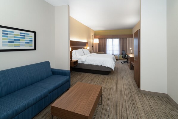 A hotel room with a bed, a blue sofa, a wooden coffee table, a framed picture on the wall, and a desk with a chair. - Holiday Inn Express Hotel & Suites Talladega by IHG (Talladega)
