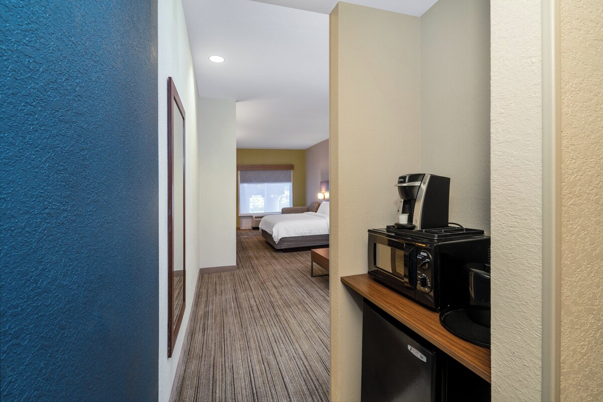 A hotel room with a bed, a desk with a coffee maker, and a view of another room with a window.