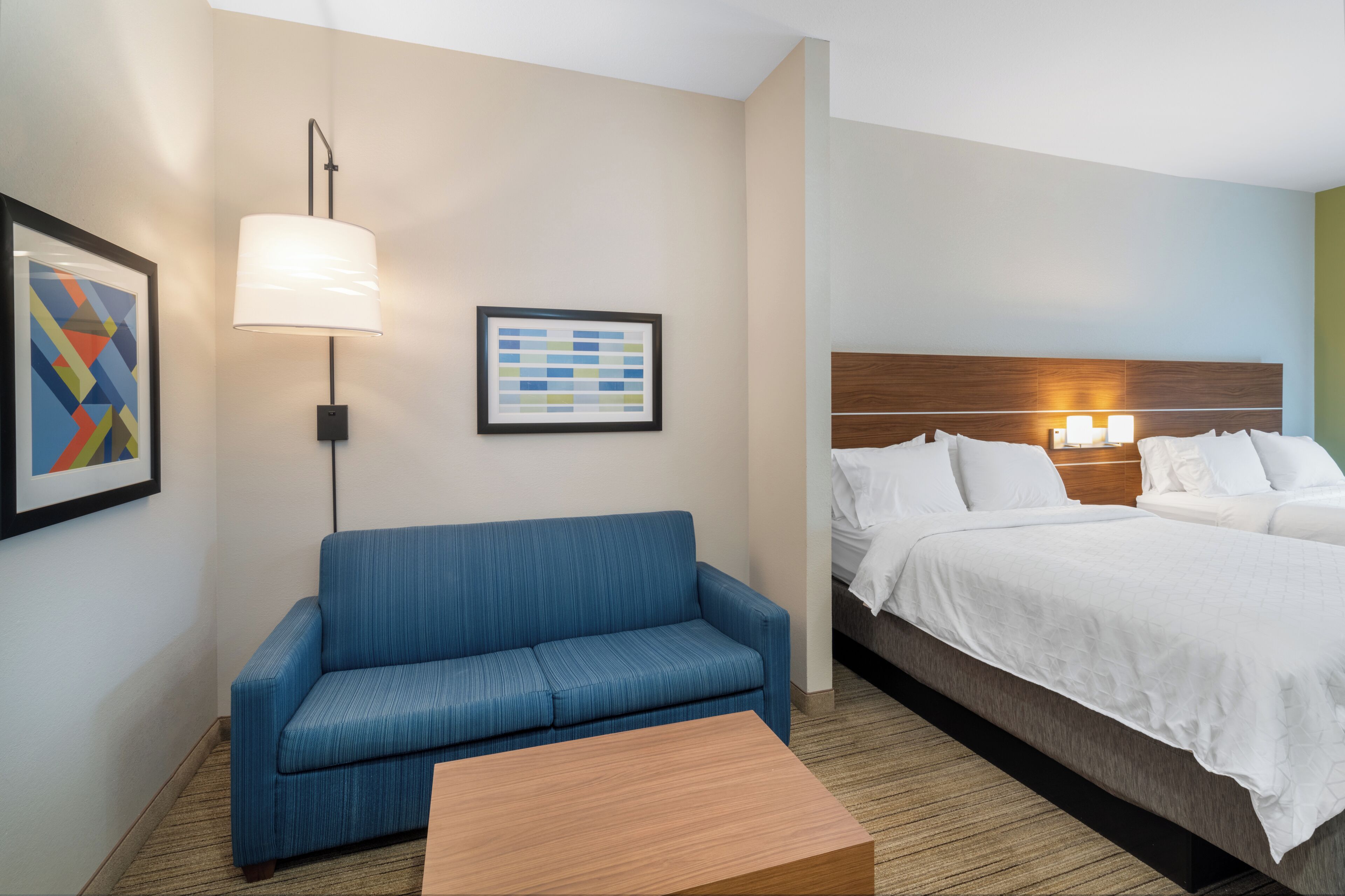 A hotel room with a blue sofa, a wooden coffee table, a bed with white bedding, a headboard with a wooden panel, and two wall-mounted lamps.