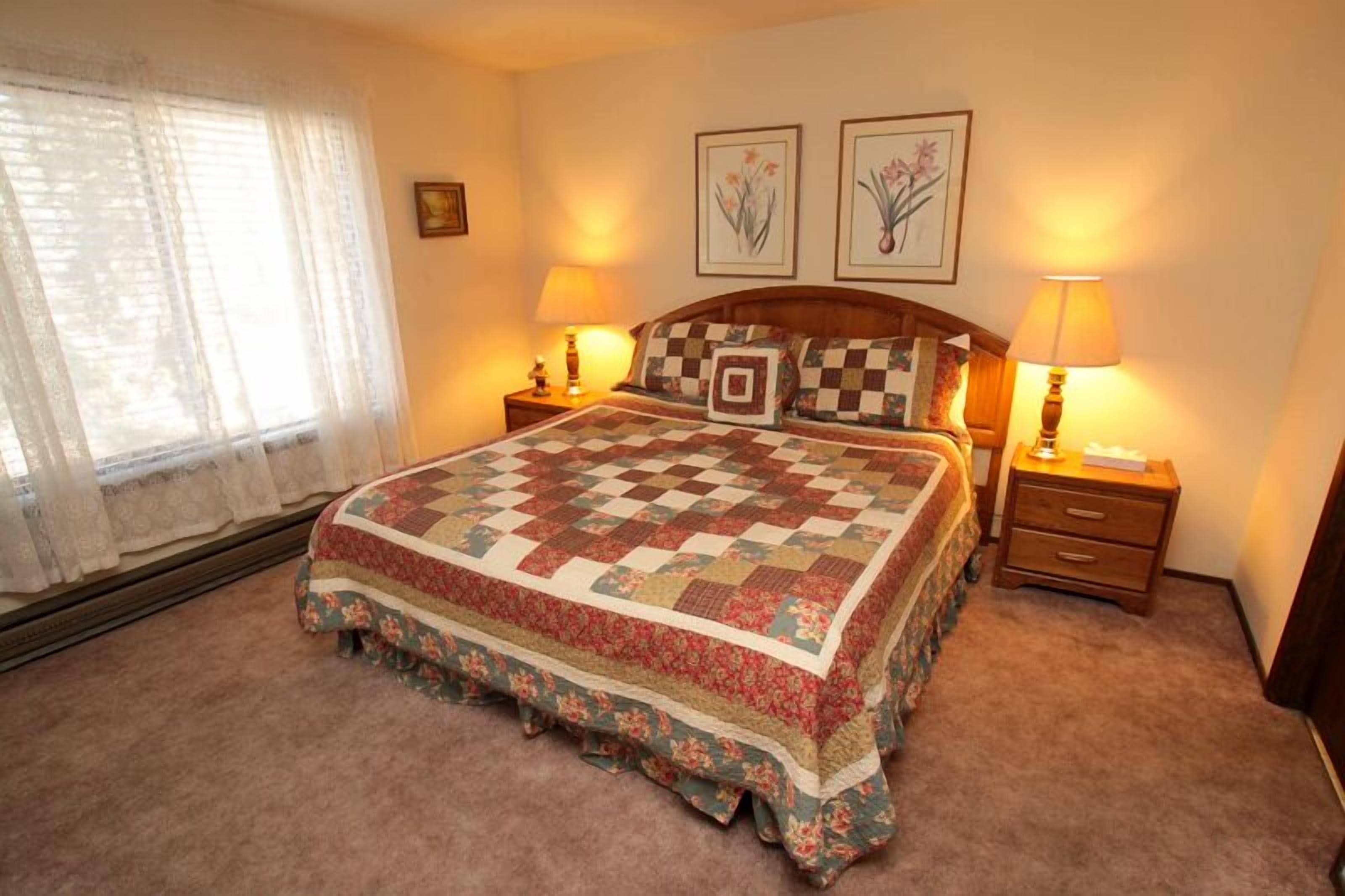 A bedroom with a large bed, floral bedspread, two bedside tables with lamps, and framed floral artwork on the wall.