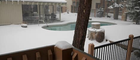 Un espacio exterior cubierto de nieve con piscina, terraza de madera y un edificio con grandes ventanales.