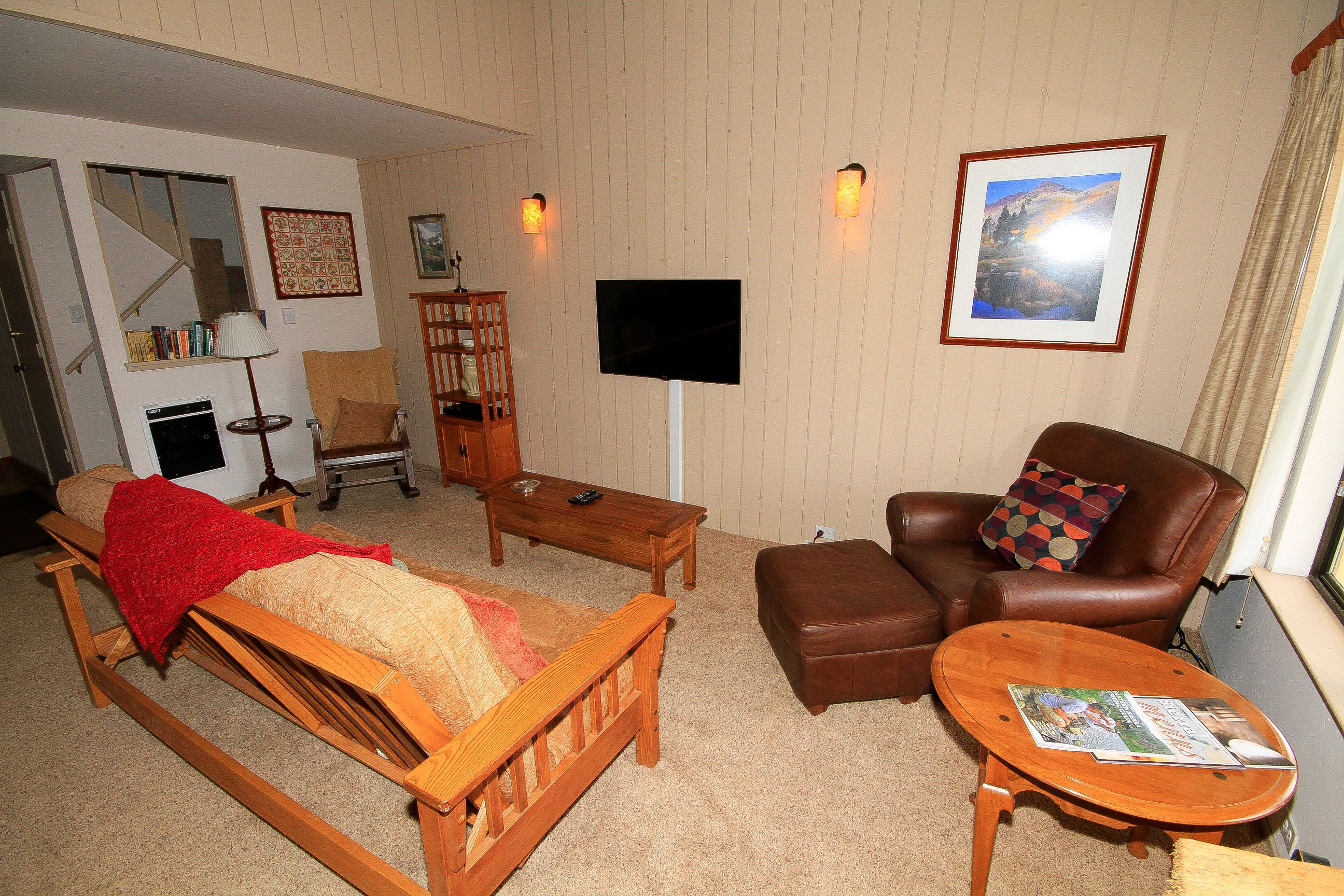 A living room with a wooden sofa, a brown leather armchair, a wooden coffee table, and a flat-screen TV mounted on the wall.