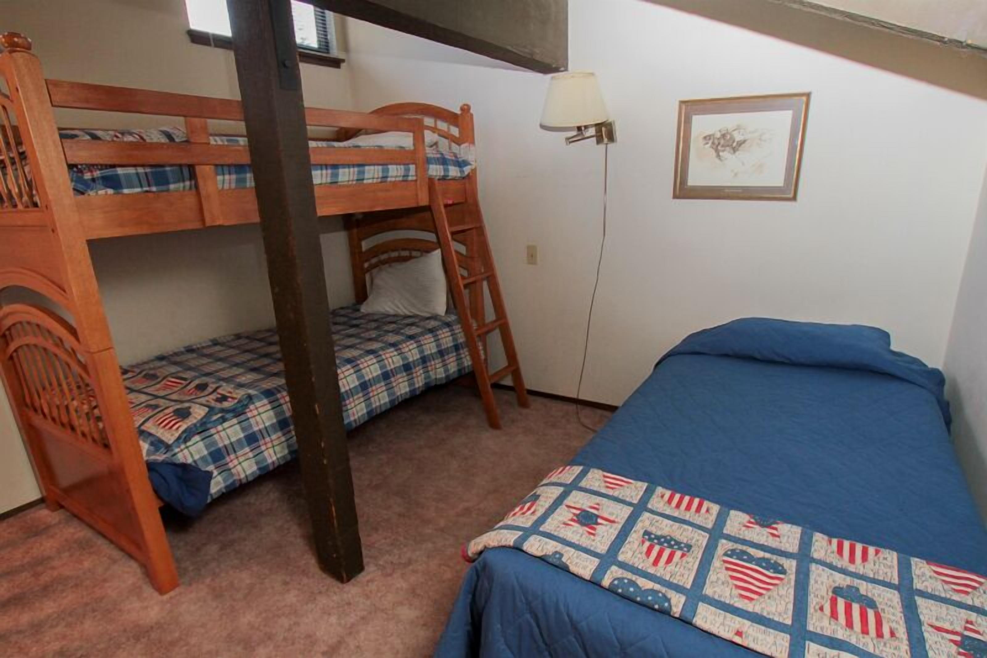 Quarto com beliche, uma cama de solteiro, escada, abajur e um quadro na parede.