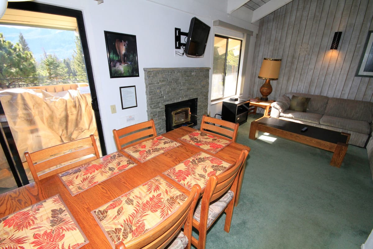 Un área de comedor con una mesa y sillas de madera, una chimenea, un sofá y un televisor montado en la pared.