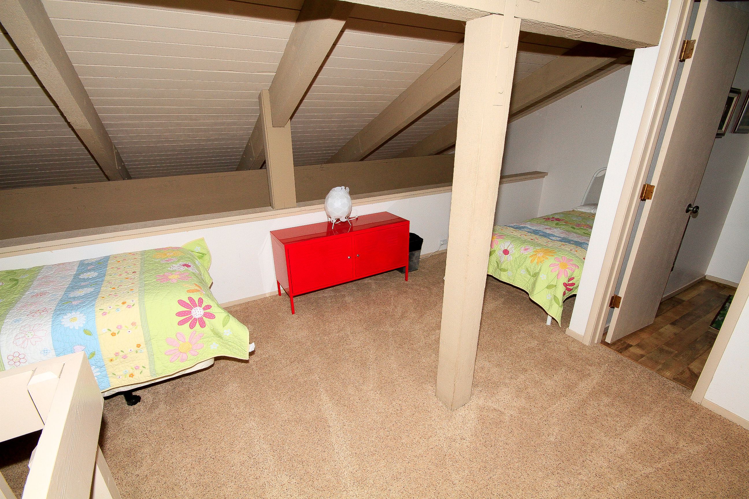 Une chambre avec un plafond en pente, une commode rouge, deux lits avec une literie à motifs floraux et une chaise en bois.