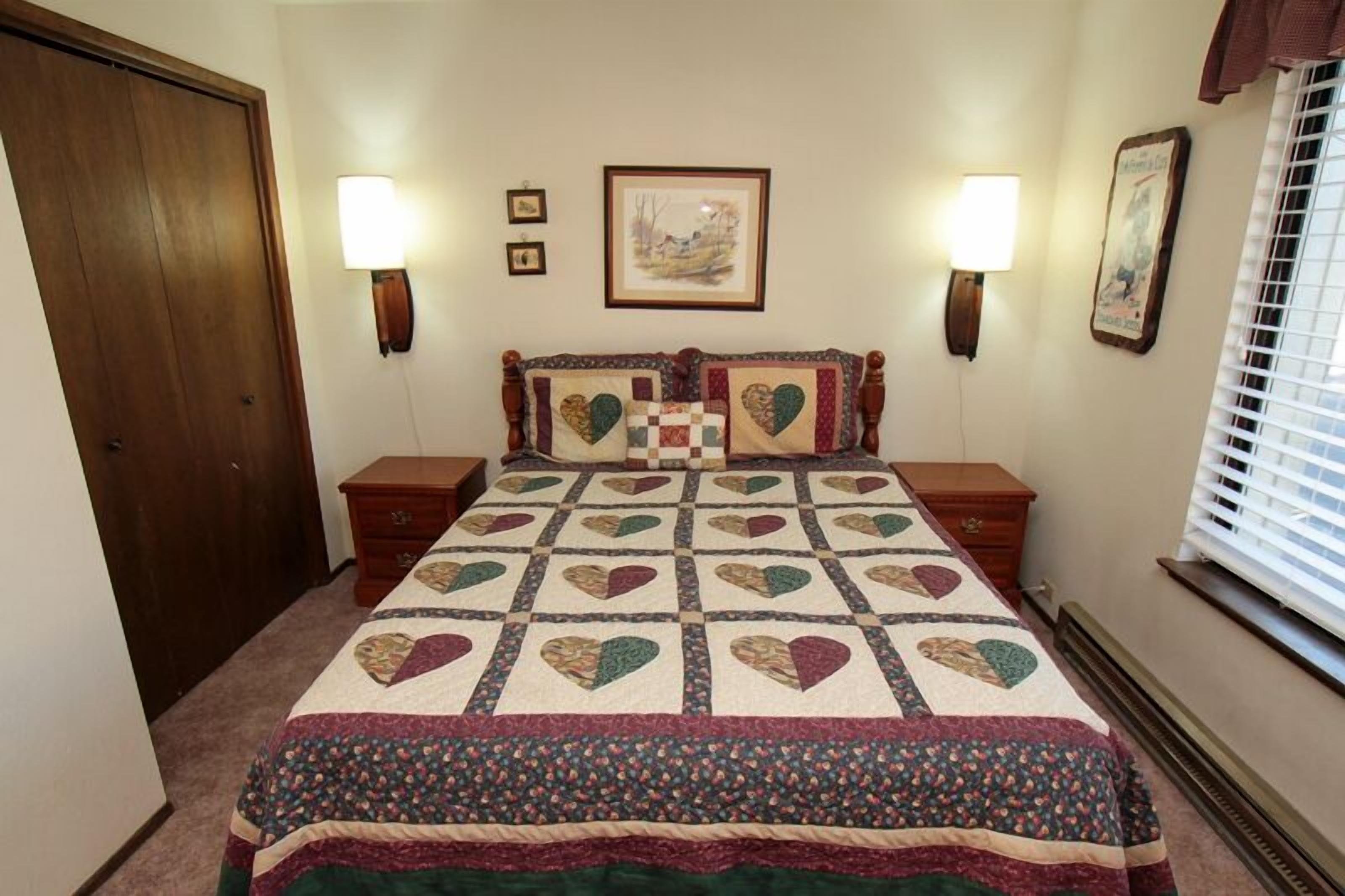 A bedroom with a large bed, two bedside tables, a wooden wardrobe, and a window with blinds.