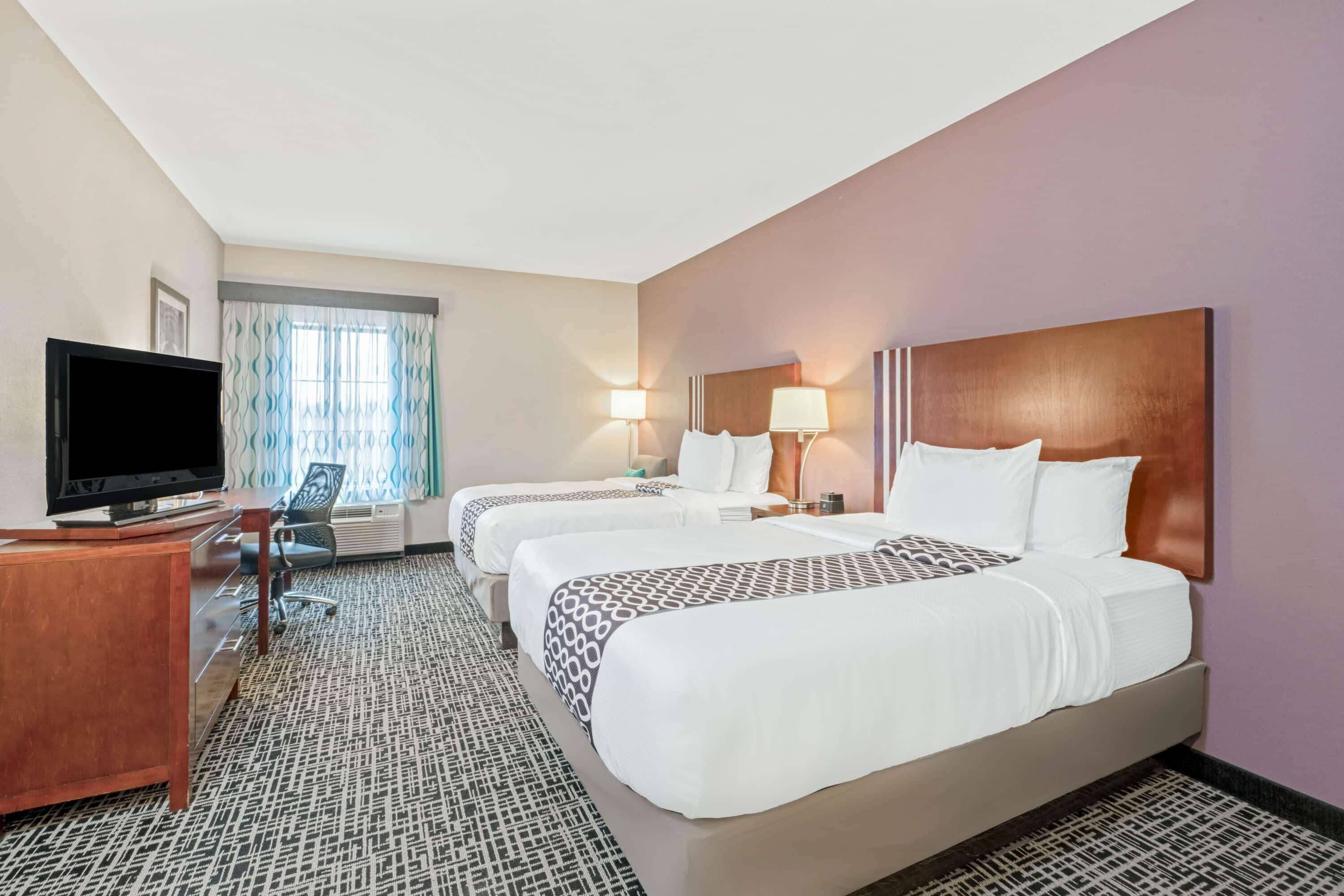 A hotel room with two beds, a desk with a flat-screen TV, a chair, and a window with curtains.