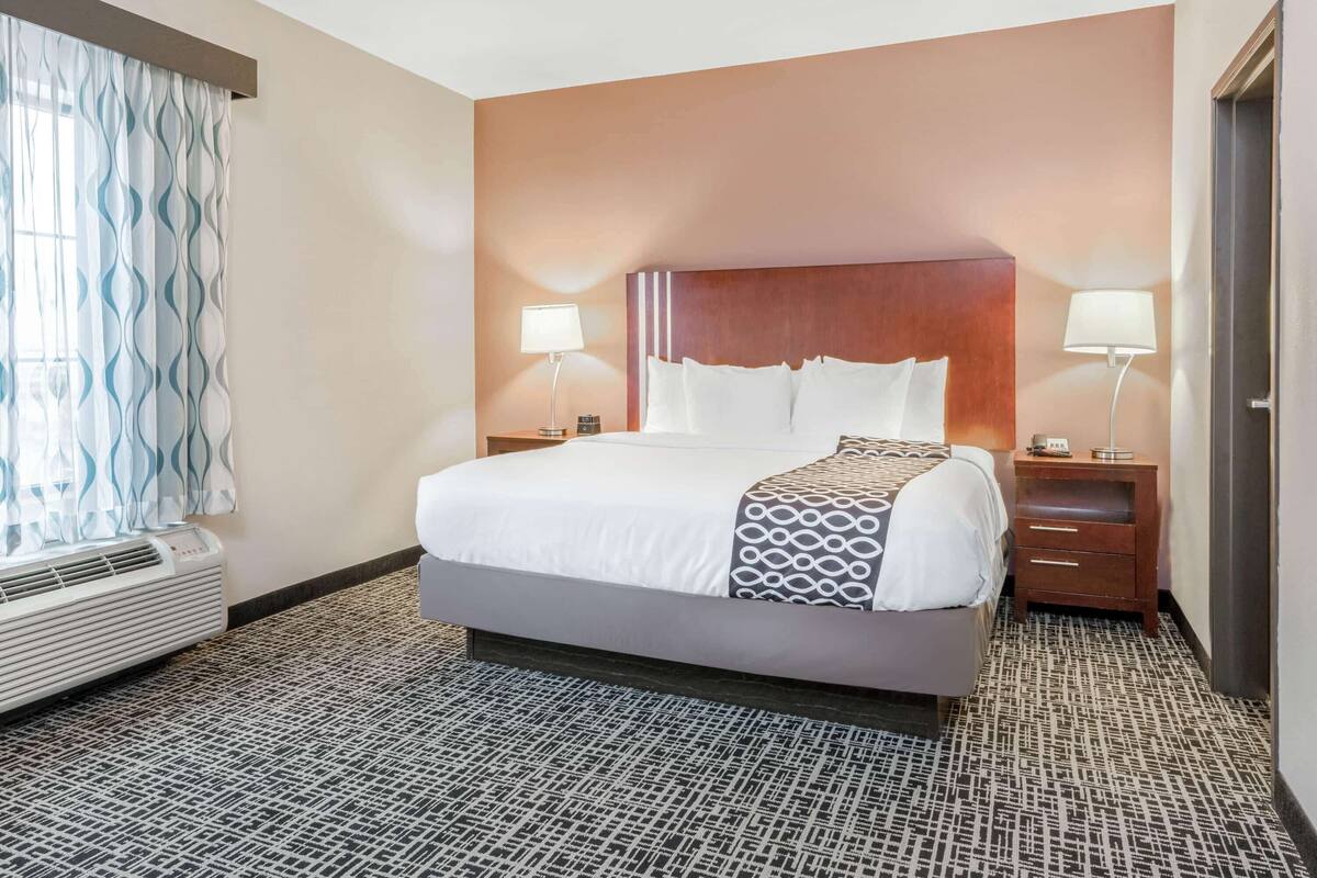 A hotel room with a large bed, two bedside tables with lamps, a window with curtains, and a patterned carpet.