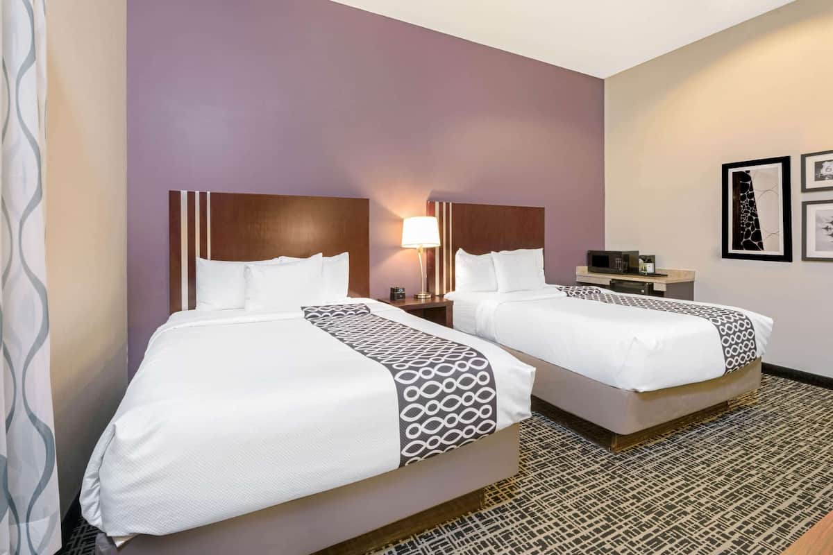 A hotel room with two beds, a nightstand with a lamp, a small table with a microwave, and framed pictures on the wall.