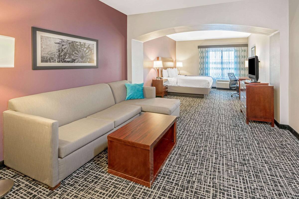 A hotel room with a sofa, a wooden coffee table, a bed, a TV, and a framed picture on the wall.