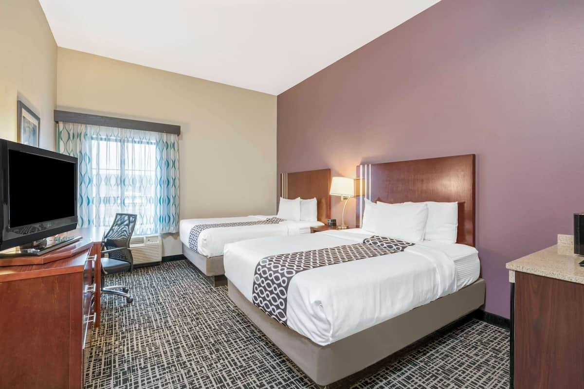 A hotel room with two beds, a desk with a flat-screen TV, a chair, and a window with patterned curtains.