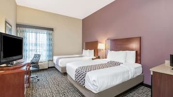 A hotel room with two beds, a desk with a flat-screen TV, a chair, and a window with patterned curtains.