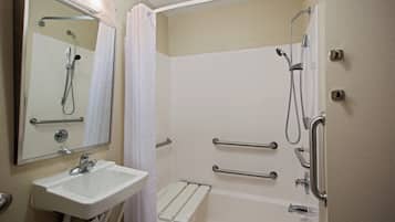 Baño con lavamanos blanco, espejo, ducha con cabezal de ducha de mano y barras de apoyo.