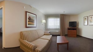 Quarto de hotel com sofá listrado, mesa de centro de madeira, TV de tela plana em suporte de madeira, quadro pendurado na parede e janela com cortinas.