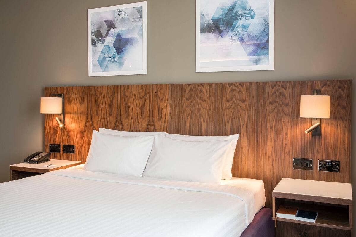 Una habitación de hotel con cabecera de madera, dos lámparas de noche, un teléfono, una mesita de noche con un libro y arte abstracto en las paredes.