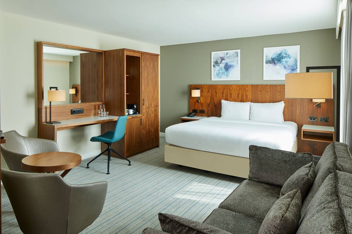 Habitación de hotel con cama, escritorio con silla, sofá, armario y dos lámparas fijadas a la pared.