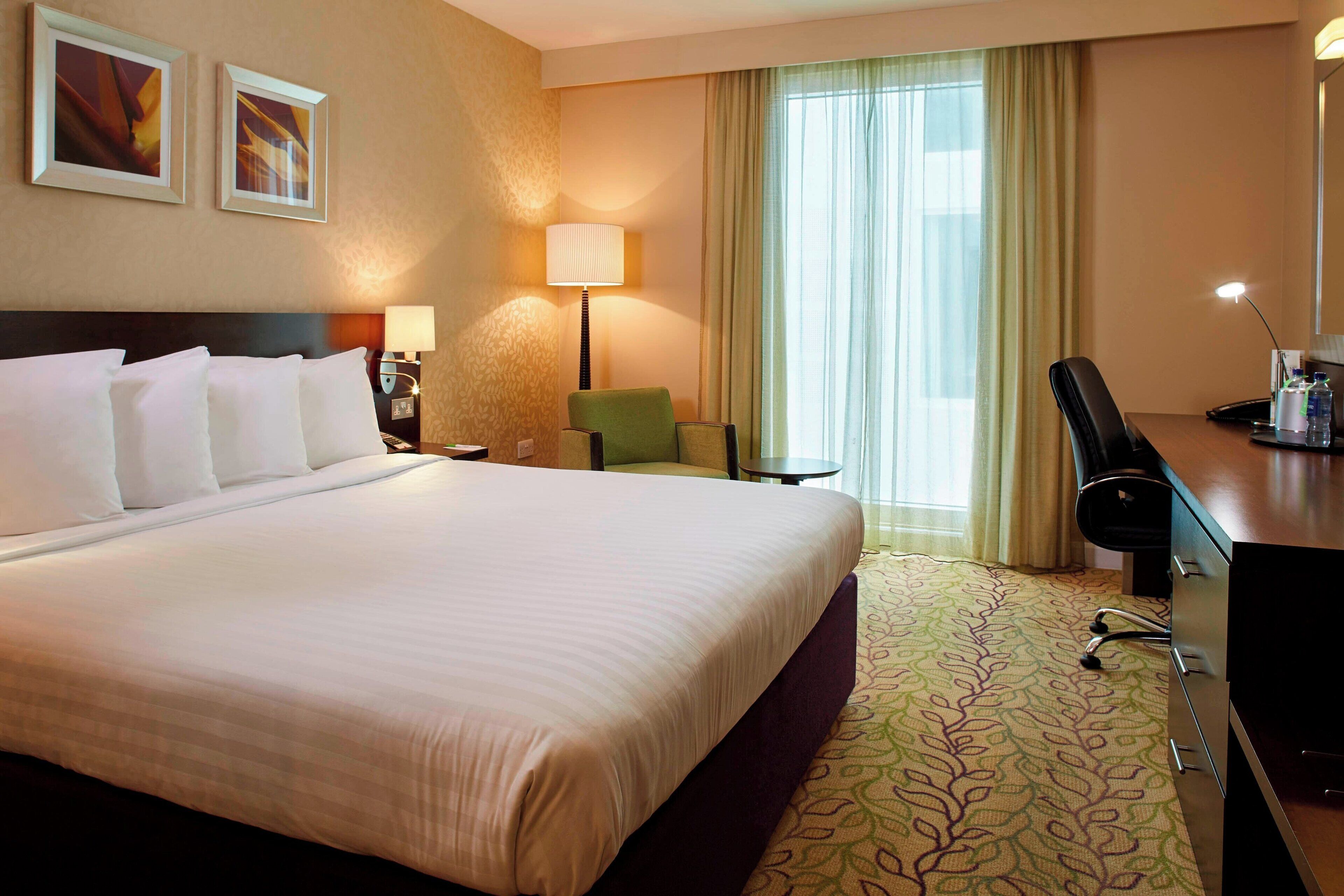 A hotel room with a large bed, a desk with a chair, a lamp, and a window with curtains.