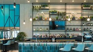 Un espacio moderno de bar con una barra de color aguamarina, varias estanterías llenas de botellas, plantas colgantes y sillas azules.