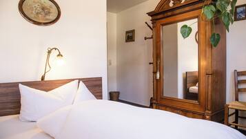 A neatly made bed with white linens and pillows, a wooden wardrobe with a mirror, a wicker basket, and a wall-mounted lamp.