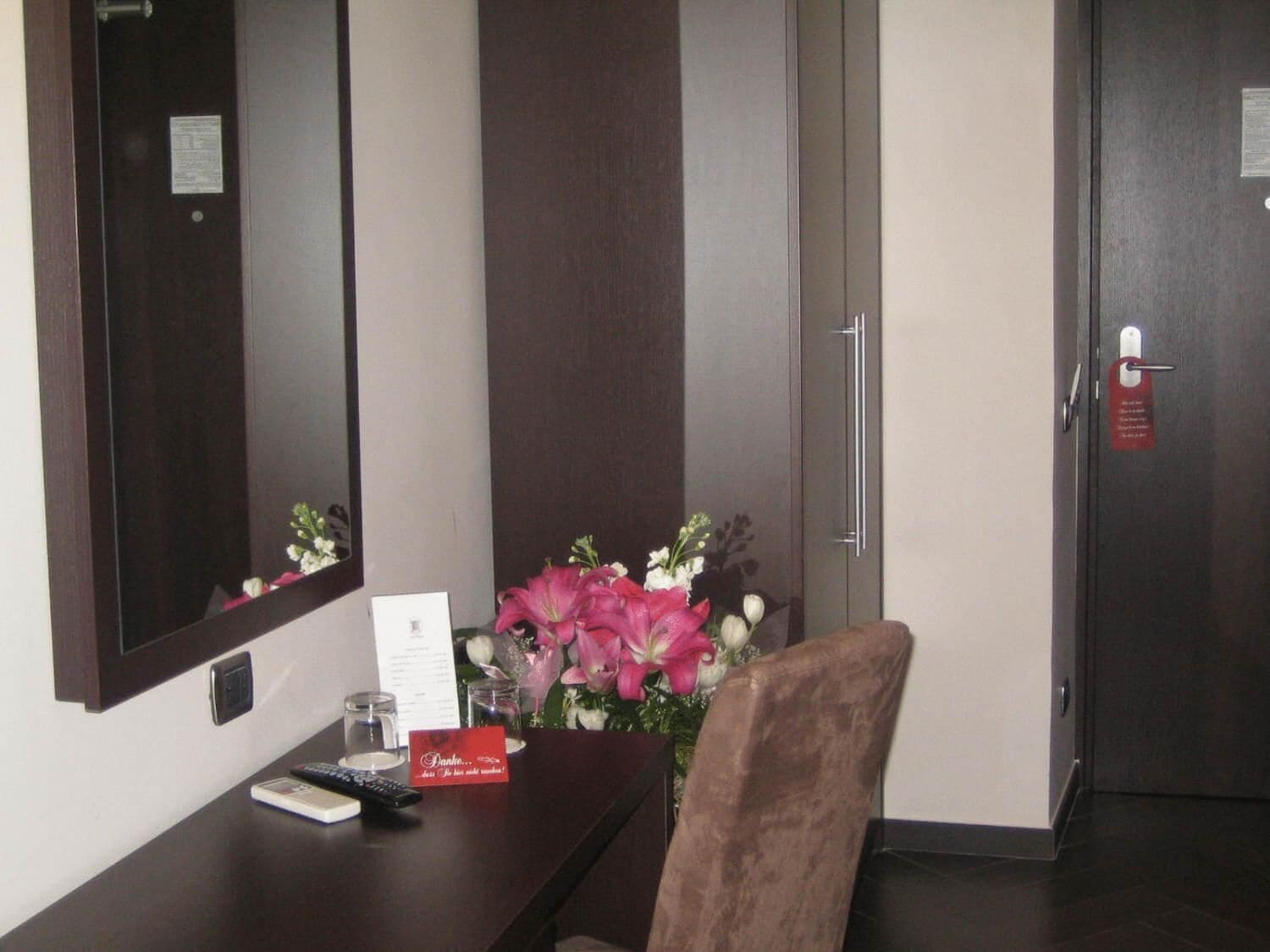 Une chambre d’hôtel avec un bureau en bois foncé, un miroir, un vase avec des fleurs roses, une télécommande et une carte rouge.