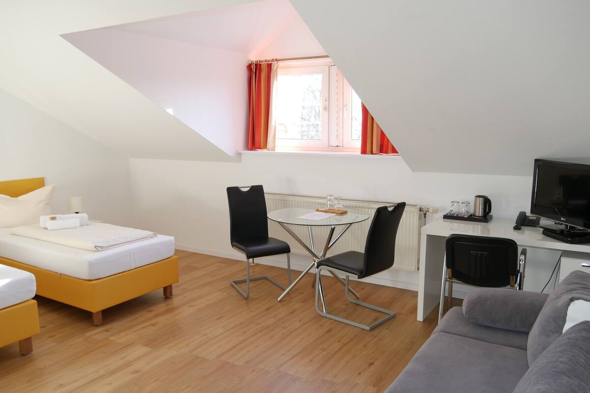 A modern, compact room with a bed, a small dining table with chairs, a sofa, a TV, and a window with curtains.