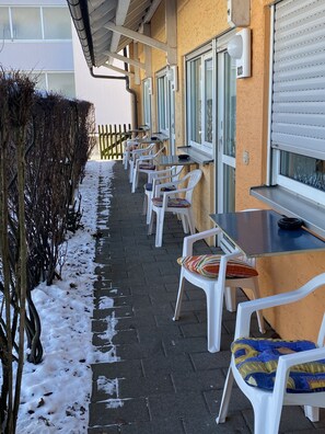 Une rangée de chaises et de tables en plastique blanc est disposée sur un trottoir pavé, et de la neige recouvre le sol.