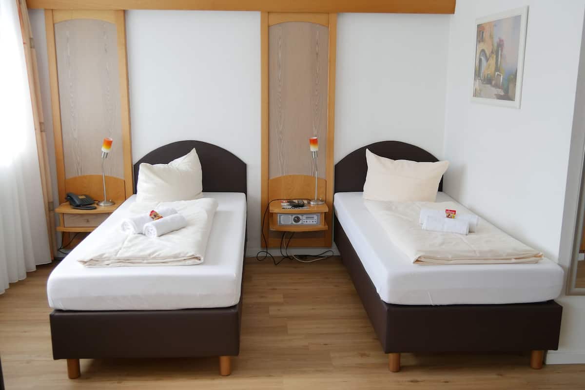 A hotel room with two single beds, wooden headboards, bedside tables with lamps, and a painting on the wall.