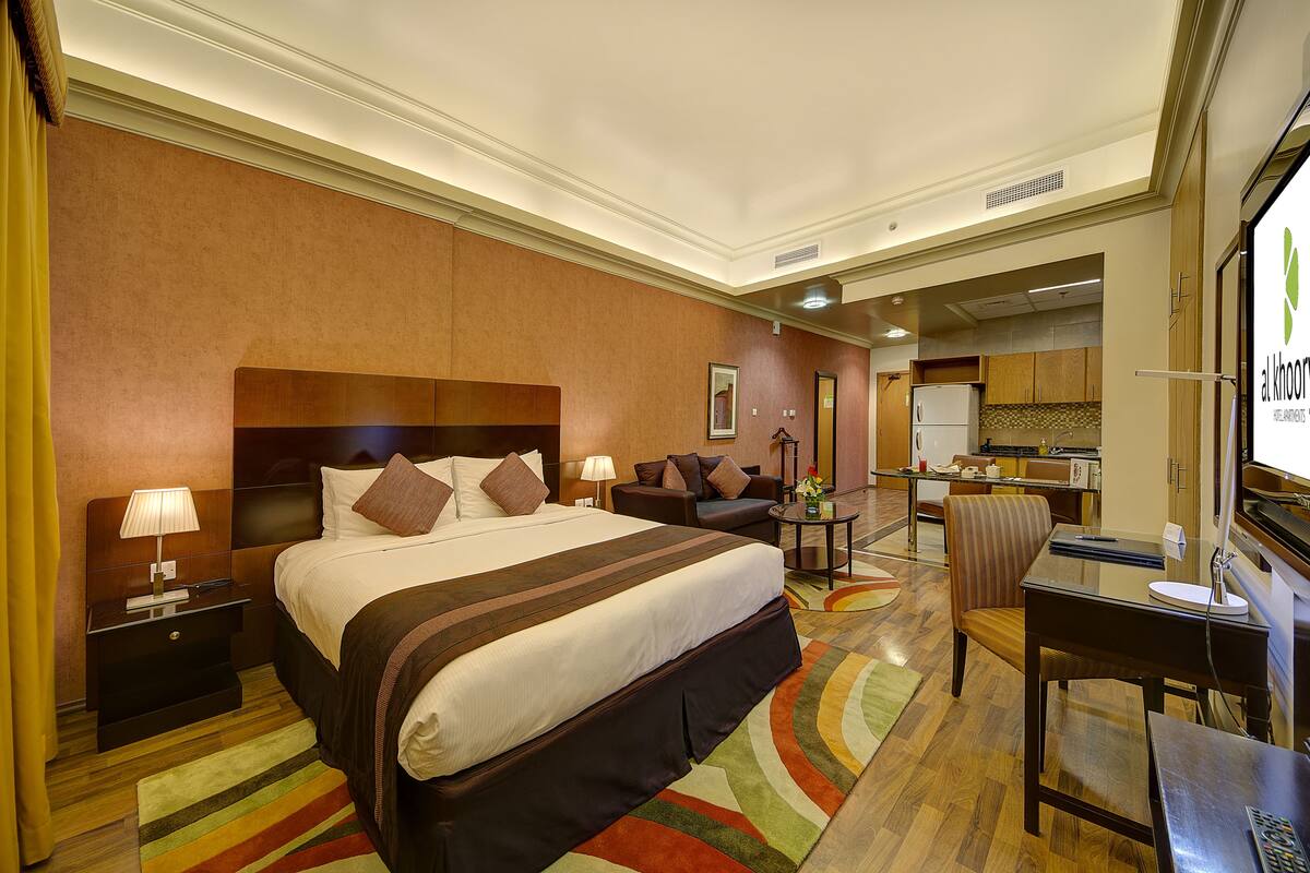 A hotel room with a large bed, a desk, a chair, a nightstand, a lamp, and a view of the kitchen area.