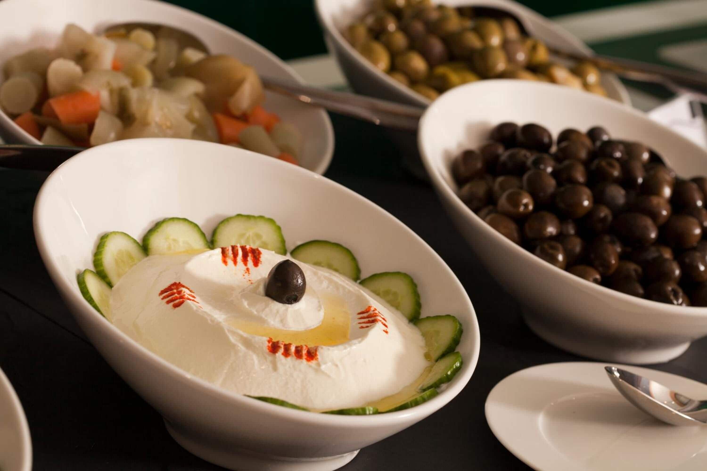 Ein Tisch mit verschiedenen Speisen, darunter eine Schüssel mit weißer Kost und Gurken sowie eine schwarze Olive, und weitere Schüsseln mit Oliven und Gemüse.