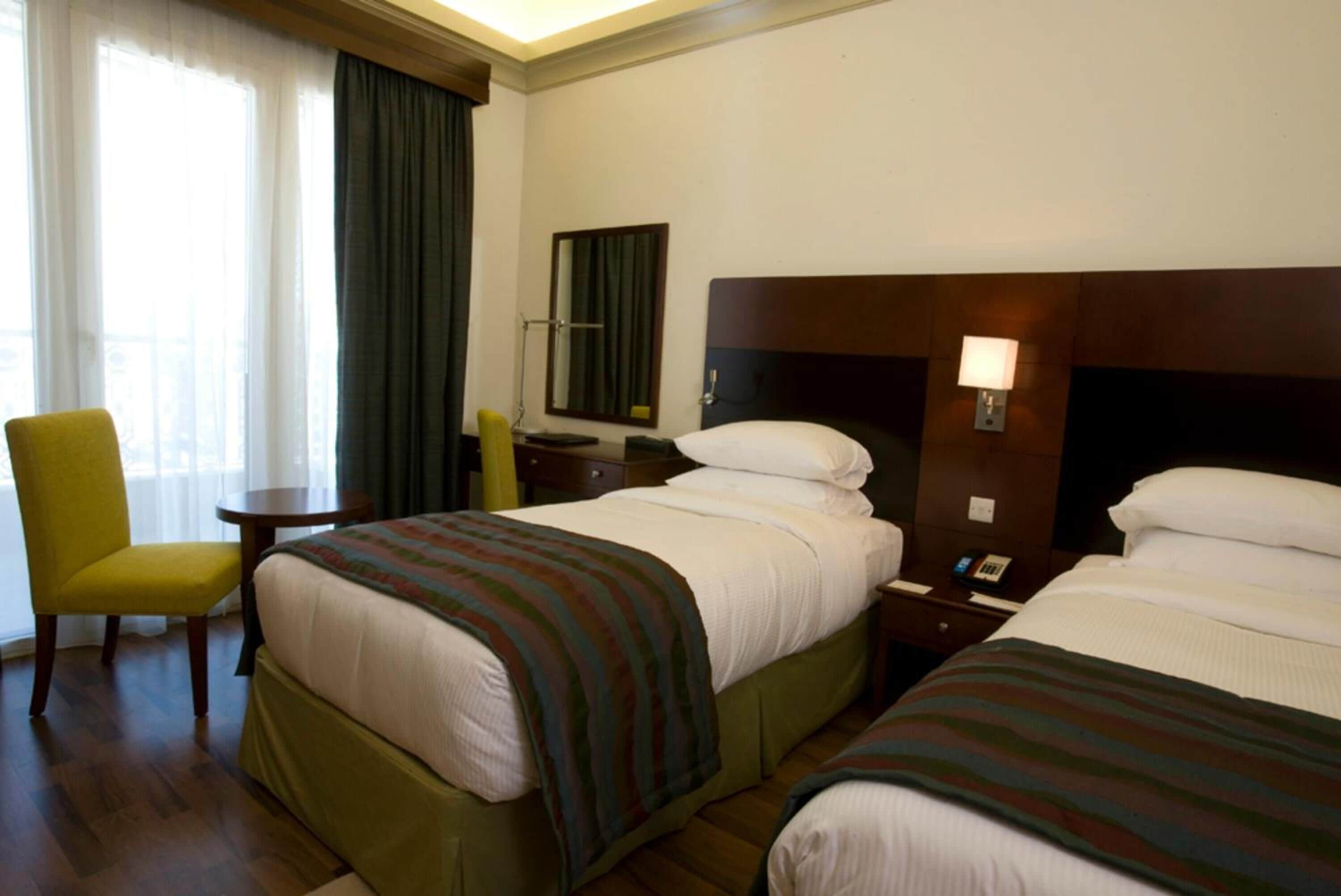 Hotellihuone, jossa on kaksi sänkyä, tuoli, työpöytä, lamppu ja ikkuna, jossa on verhot.
