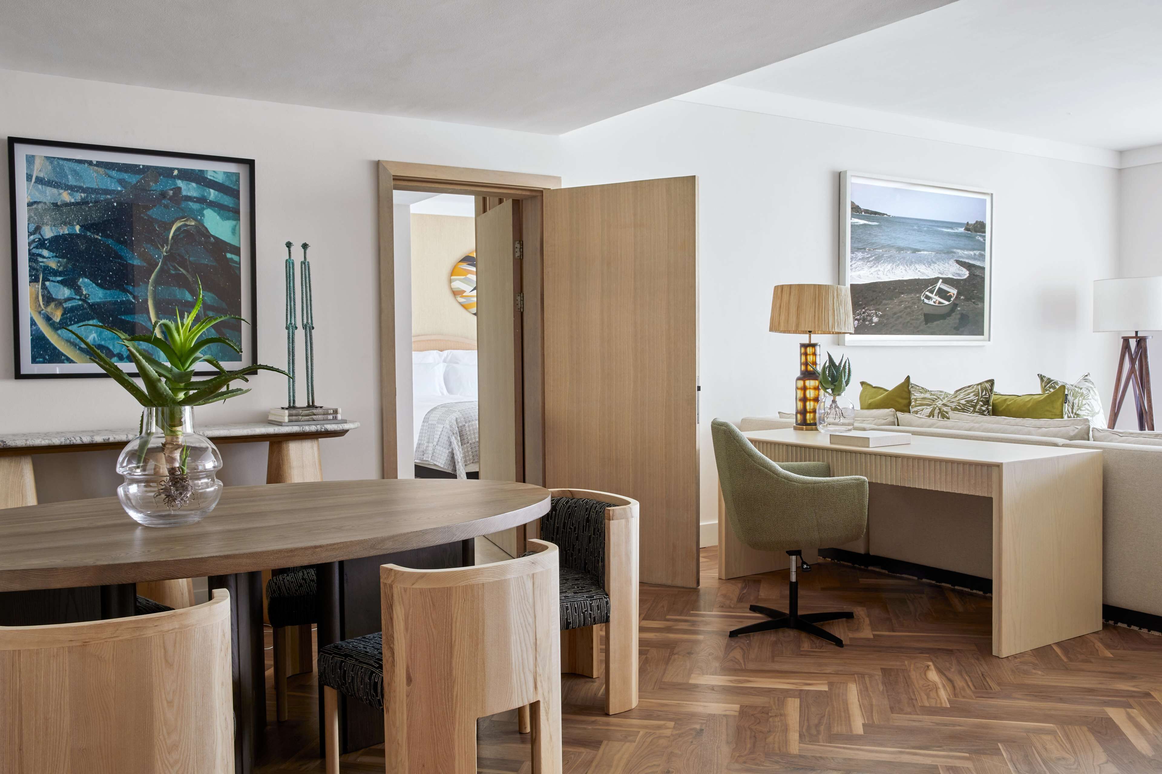 A modern hotel room with a wooden dining table, a green chair, a desk with a lamp, and a bed in the background.